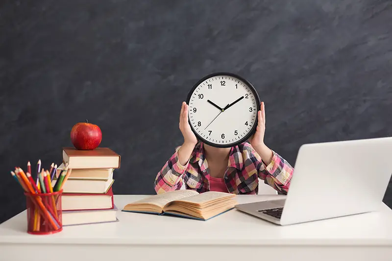Girl sitting with stack of books and laptop, holding big clock, covering face