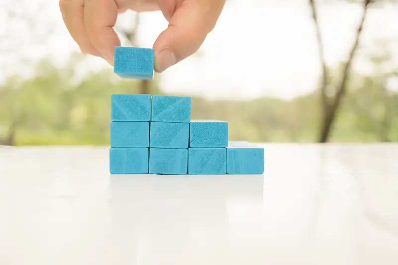 Man's hand holding blue wooden block on white wooden table.