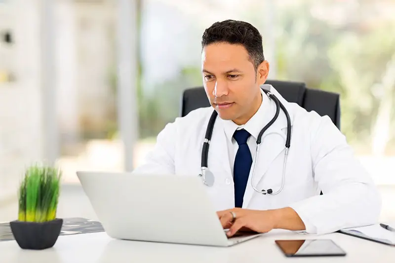 Doctor or medical consultant in white coat with and stethoscope sitting at a desk working on laptop