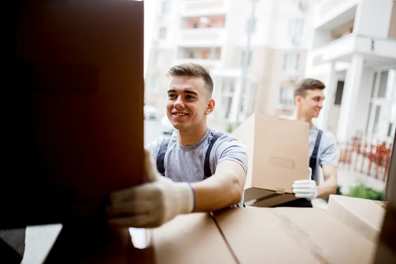 A young handsome smiling mover wearing uniform is reaching for to grab a box in removal van