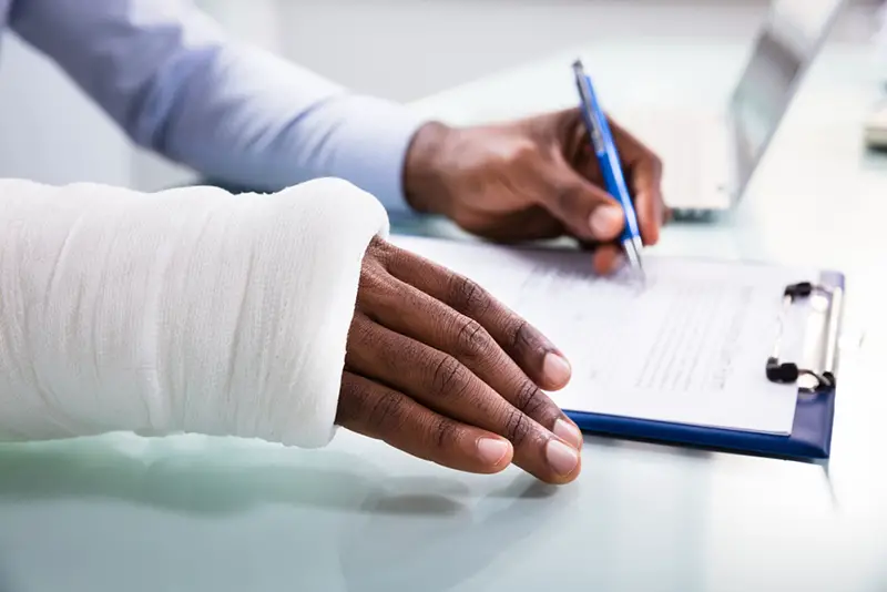 Overhead View Of Injured Man With Bandage Hand Filling Insurance Claim Form On Clipboard