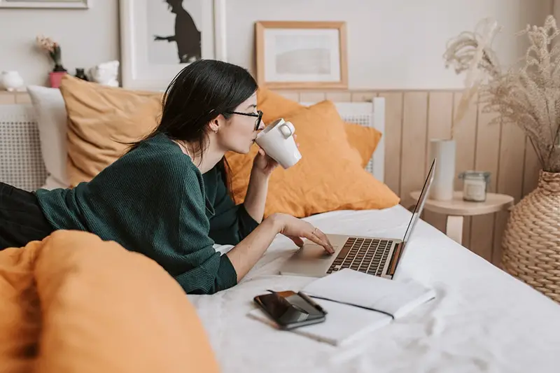 Young woman wearing eyeglasses drinking coffee in front of her laptop in bed