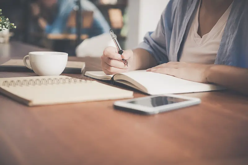 woman writing on a notebook beside a teacup and tablet computer