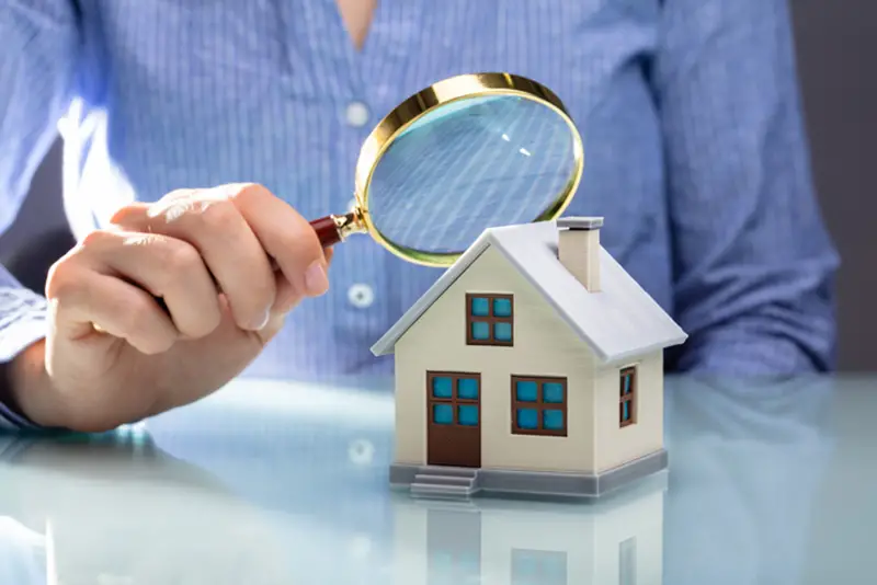 Close-up Of A Businesswoman's Hand Holding Magnifying Glass Over House Model Over Desk