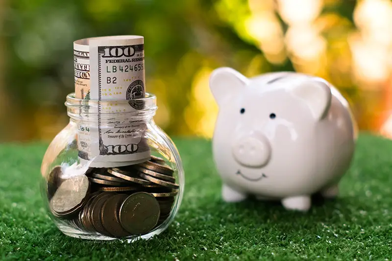 Money inside glass jar beside white piggy bank