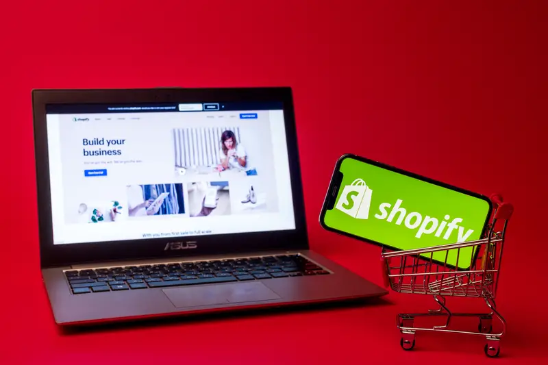 Shopify web page displayed on a modern laptop on red background. Shopify logo on iphone X in shopping cart