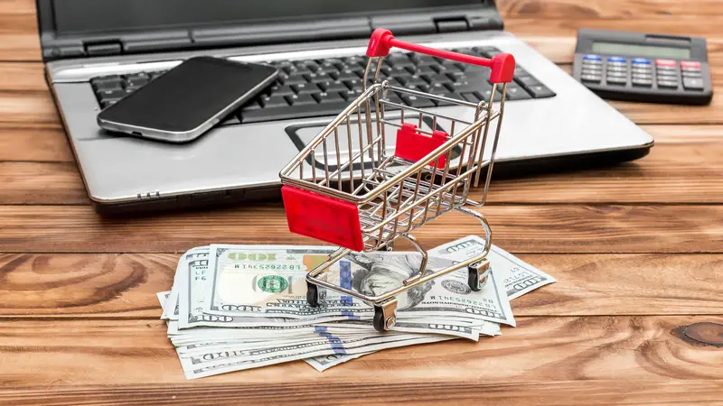 Shopping cart with money and credit card, laptop and smartphone on the table.