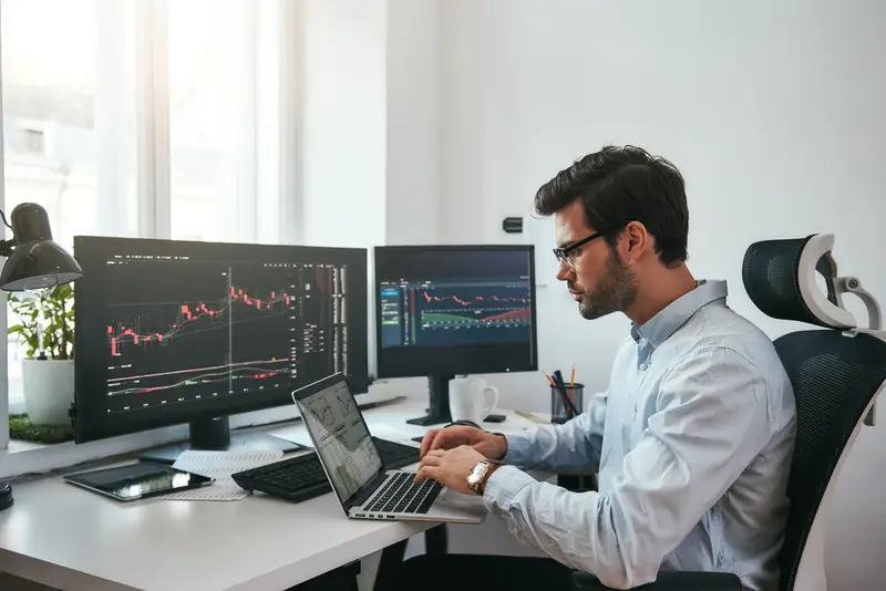 Workplace of trader. Young bearded trader wearing eyeglasses using his laptop while sitting in office in front of computer screens