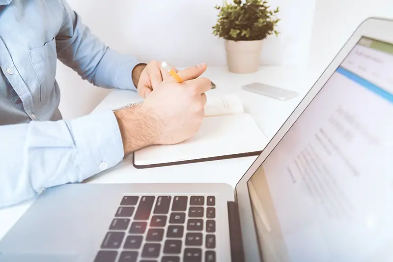 person writing on white notebook beside him a laptop