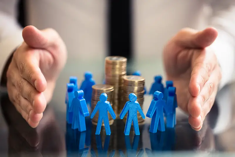 Businessperson's Hand Protecting Blue Human Figures Surrounding Stacked Golden Coins Over Desk