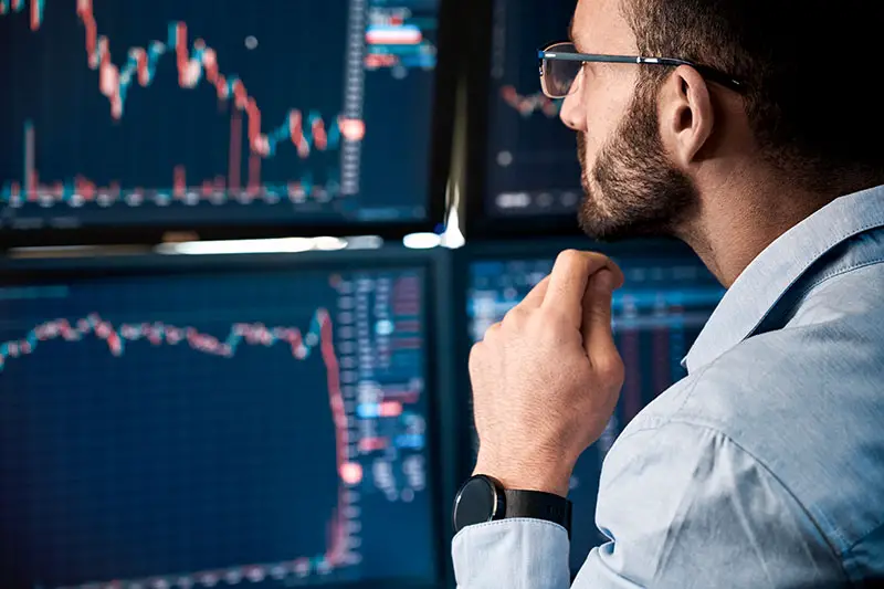 Bearded man trader wearing eyeglasses sitting at desk at office monitoring stock market