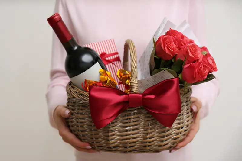 Woman holding wicker basket with gift, bouquet and wine