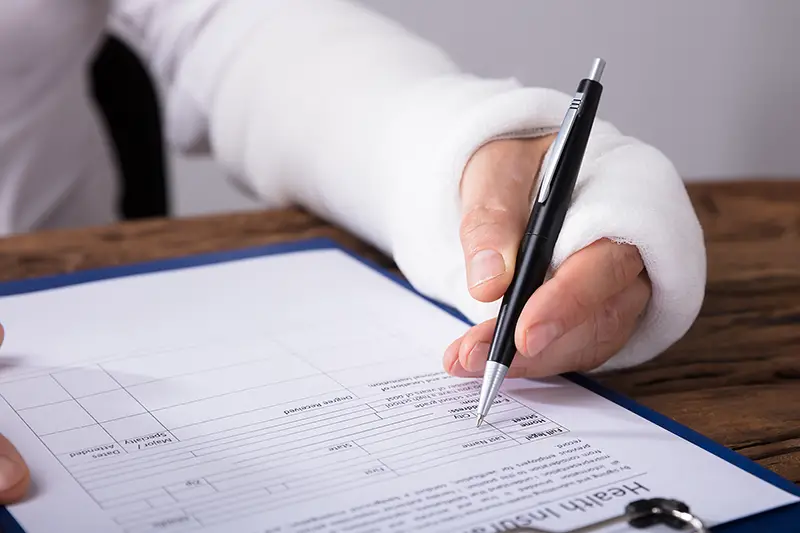 Businessperson With Broken Arm Filling Health Insurance Claim Form On Wooden Desk