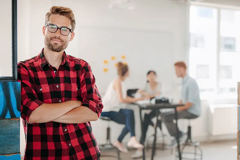 Young man standing in office with colleagues meeting in background.