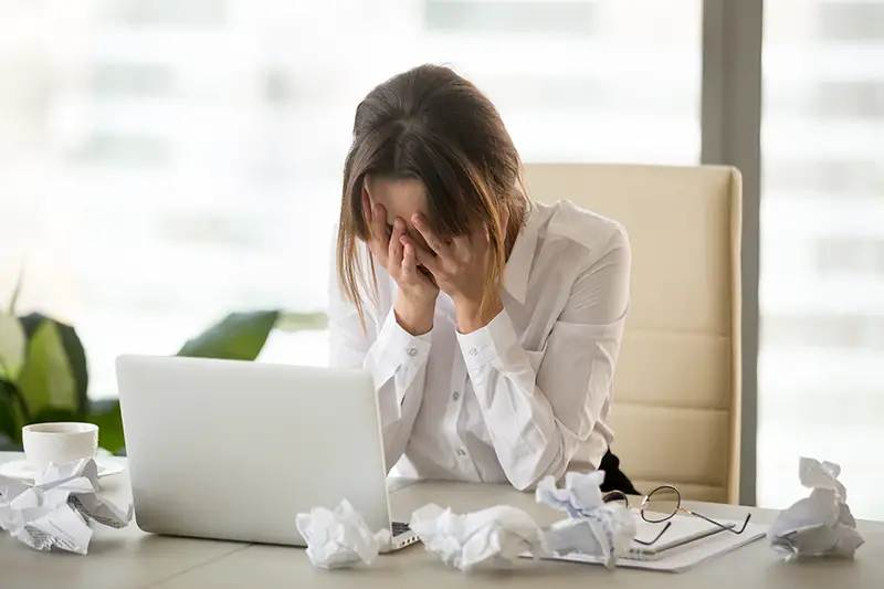 Stressed tired businesswoman feels exhausted sitting at office desk with laptop and crumpled paper