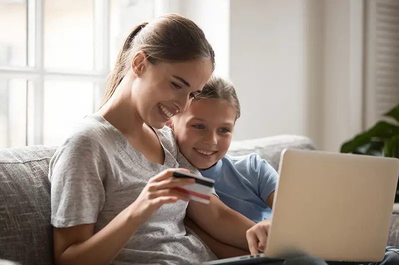 Mother and schoolchild daughter sitting on sofa in living room look at notebook screen use credit card