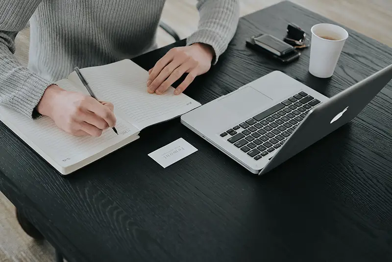 A woman takes notes from her open laptop in a notebook.