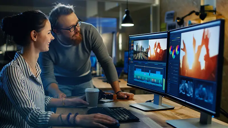 Female Video and Sound Editor Works With Her Male Colleague on a Project on Her Personal Computer with Two Displays