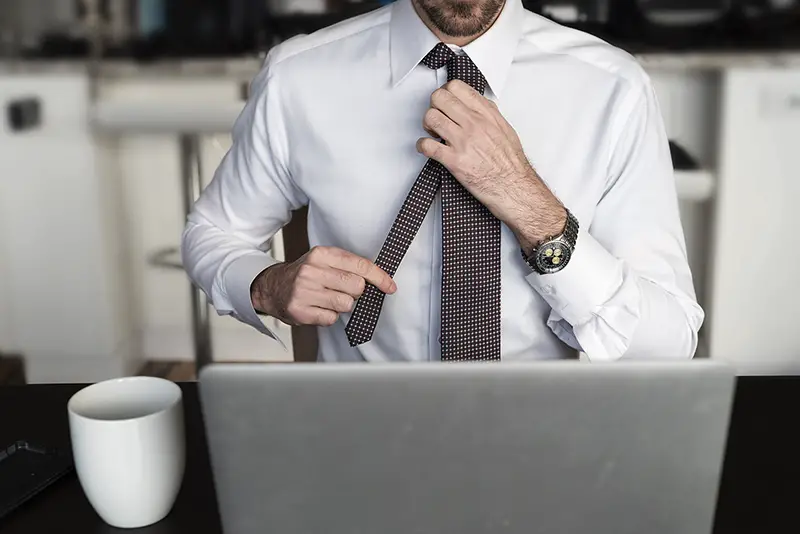 Headless man at desk adjusting his tie in front of laptop with coffee while working from home