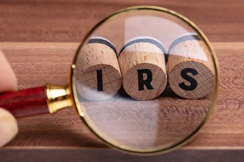 Human Hand Holding Magnifying Glass Over Wooden Cork With IRS Text On Wooden Textured