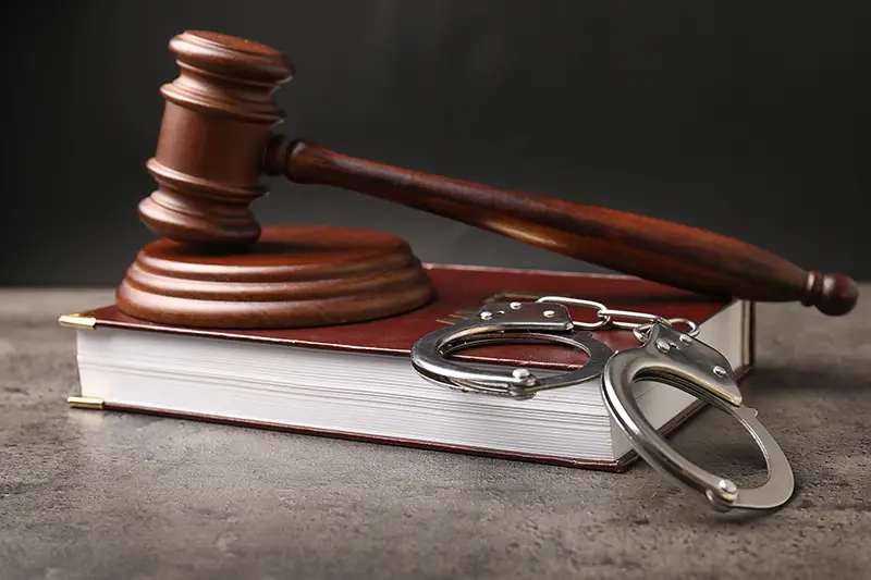 Judge's gavel, book and handcuffs on table against dark background