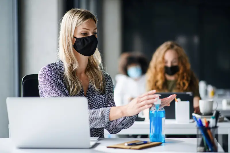 Young woman with face mask back at work in office disinfecting hands