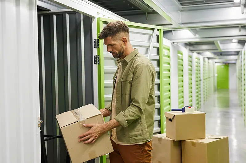 Side view at bearded adult man loading cardboard boxes into self storage unit
