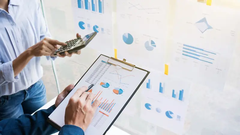 Business colleague holding a clipboard and black calculator analysing the data and chart posted on the glass wall