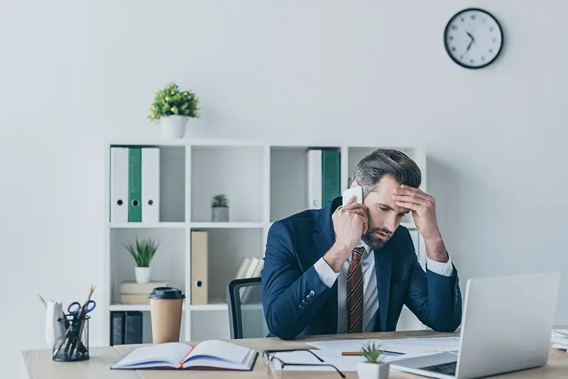 Upset businessman touching his forehead while talking on his phone at work