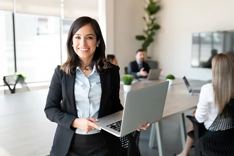 Portrait of smiling female entrepreneur holding a laptop with team in background at office conference room