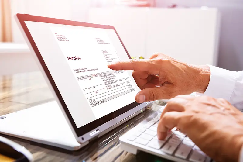 Businessperson's Hand Analyzing Invoice On Laptop At Workplace