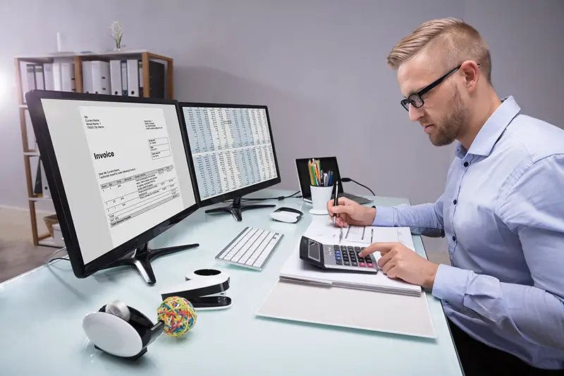 Side View Of A Young Businessman Calculating Invoice With Calculator At Workplace