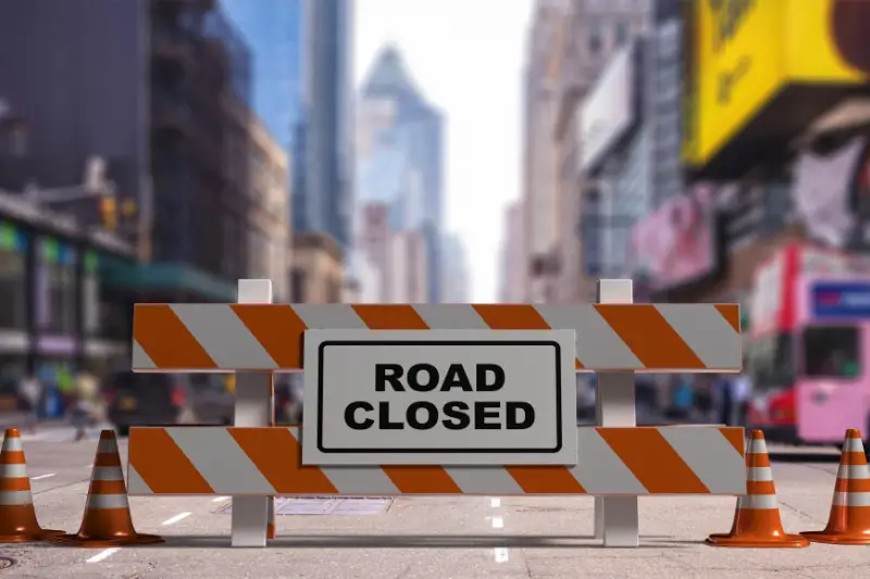 Closed road text sign, street barriers and traffic cones downtown, city center background