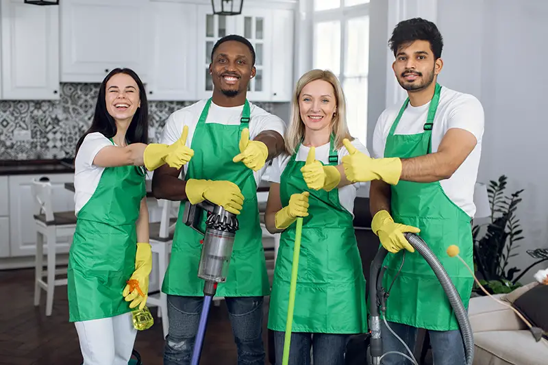 Smiling multiracial housekeepers in green aprons and yellow gloves showing thumbs up at work.