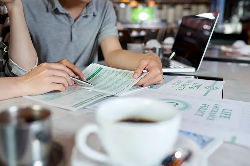 Close-up shot of unrecognizable young family sitting at cafe table and reading life insurance brochure, blurred background