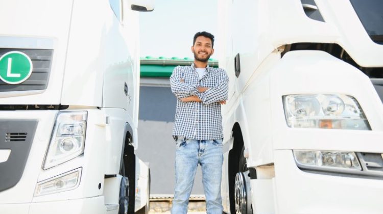 A confident Indian truck driver stood between two trucks parked in a car park.