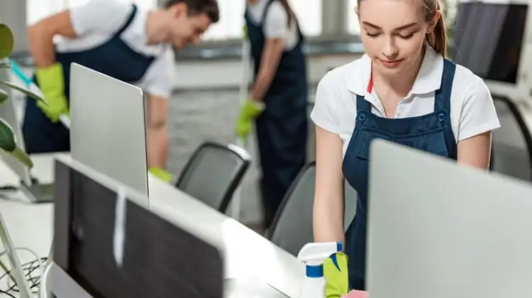 Young woman cleaner wiping desk while colleagues cleaning floor in office