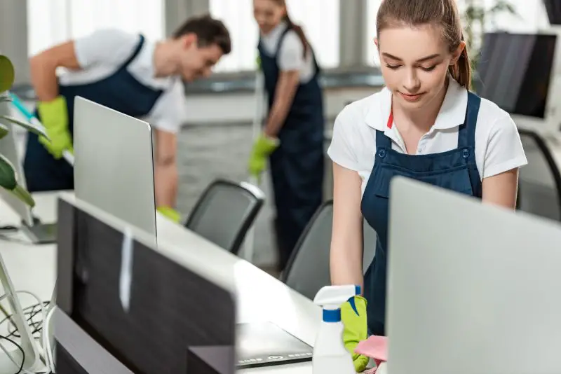 Young woman cleaner wiping desk while colleagues cleaning floor in office