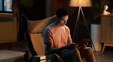Man sitting in a modern armchair under a floor lamp, using a tablet in a dimly lit, cozy room.