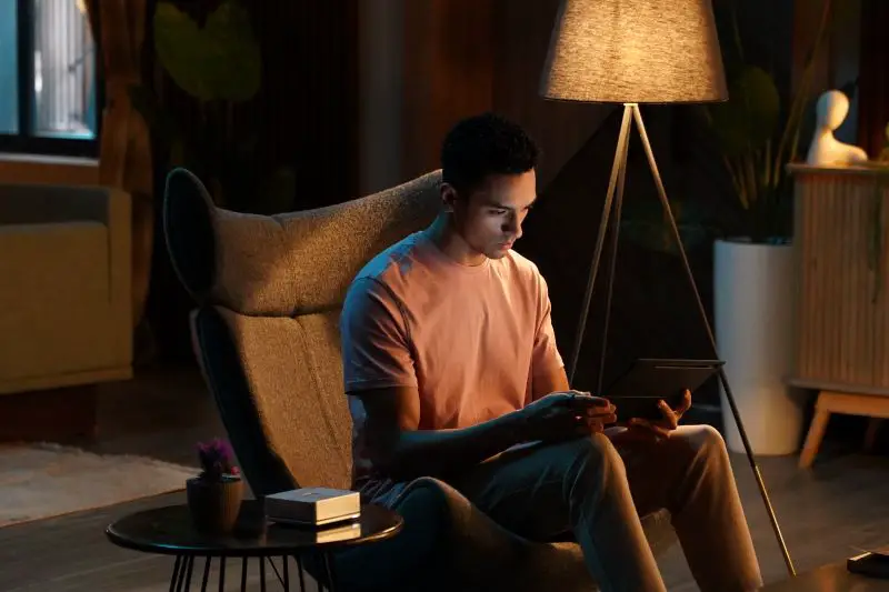 Man sitting in a modern armchair under a floor lamp, using a tablet in a dimly lit, cozy room.