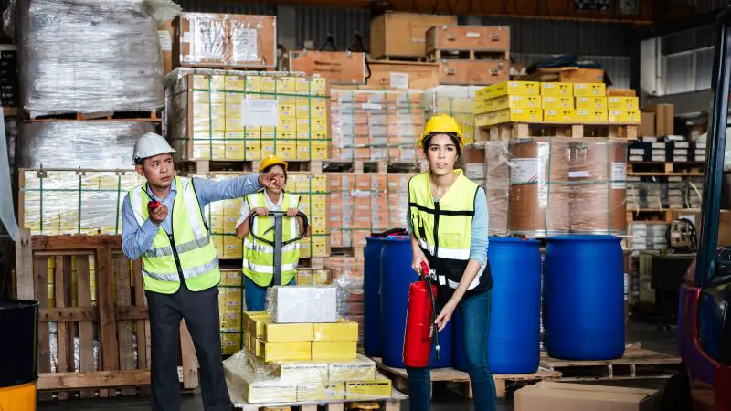 Group of warehouse workers – one with a radio, one holding a fire extinguisher, preparing to use it, and another with a pallet of goods.